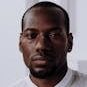 Headshot of a serious black man in a white shirt, indoors with a neutral background.